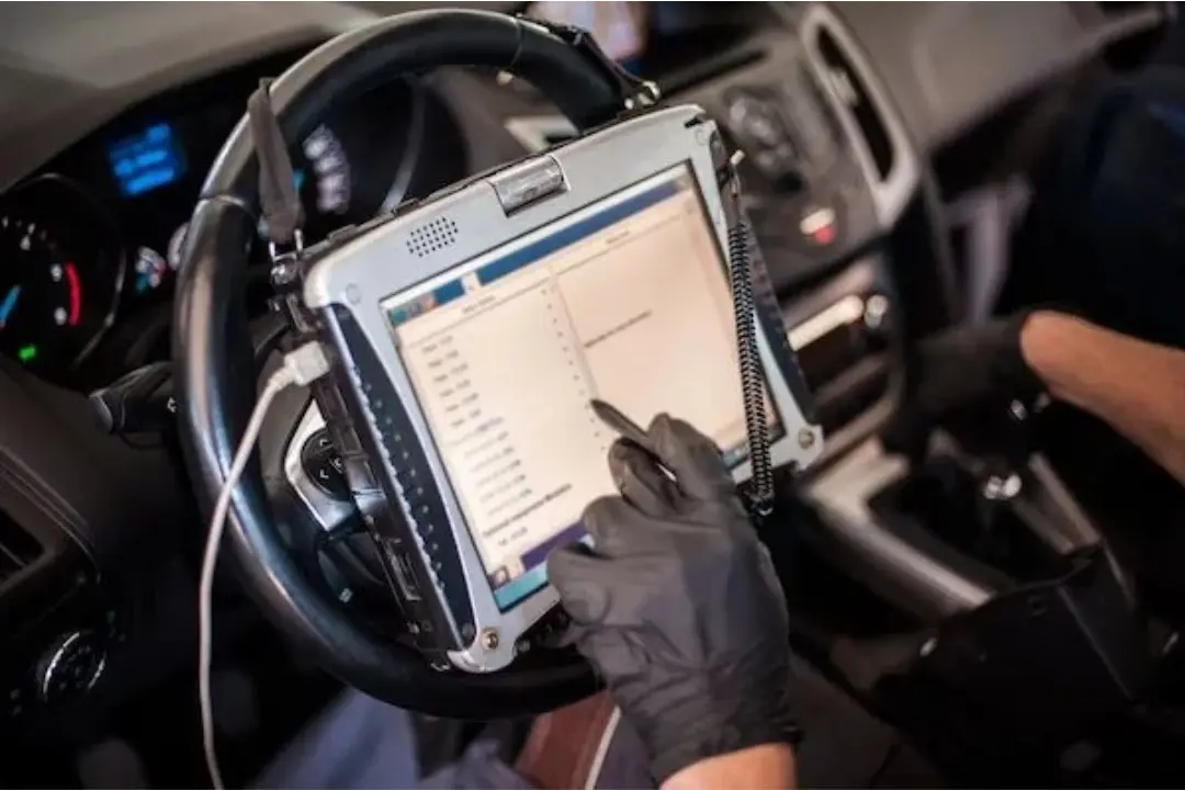 Mão com luva protetora preta manuseando uma caneta digital na tela de um tablet de diagnóstico veicular, fixado junto ao volante no interior de um automóvel.