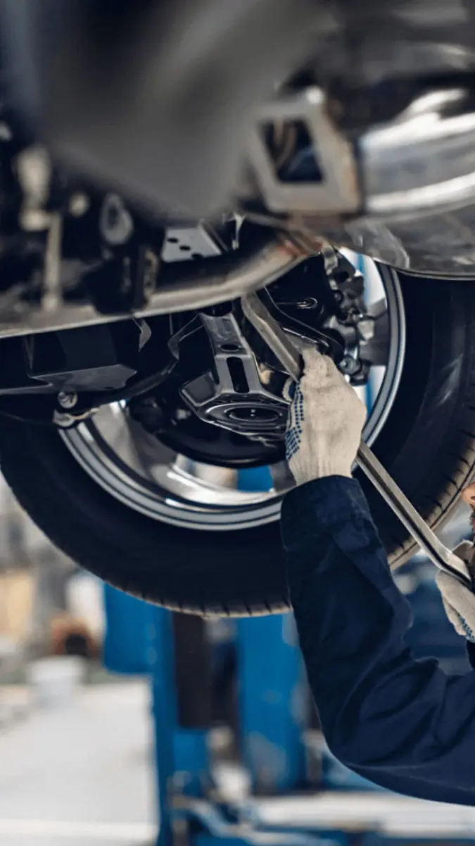 Profissional com luva protetora utiliza ferramenta mecânica para efetuar ajustes na parte inferior do chassi de um automóvel suspenso.