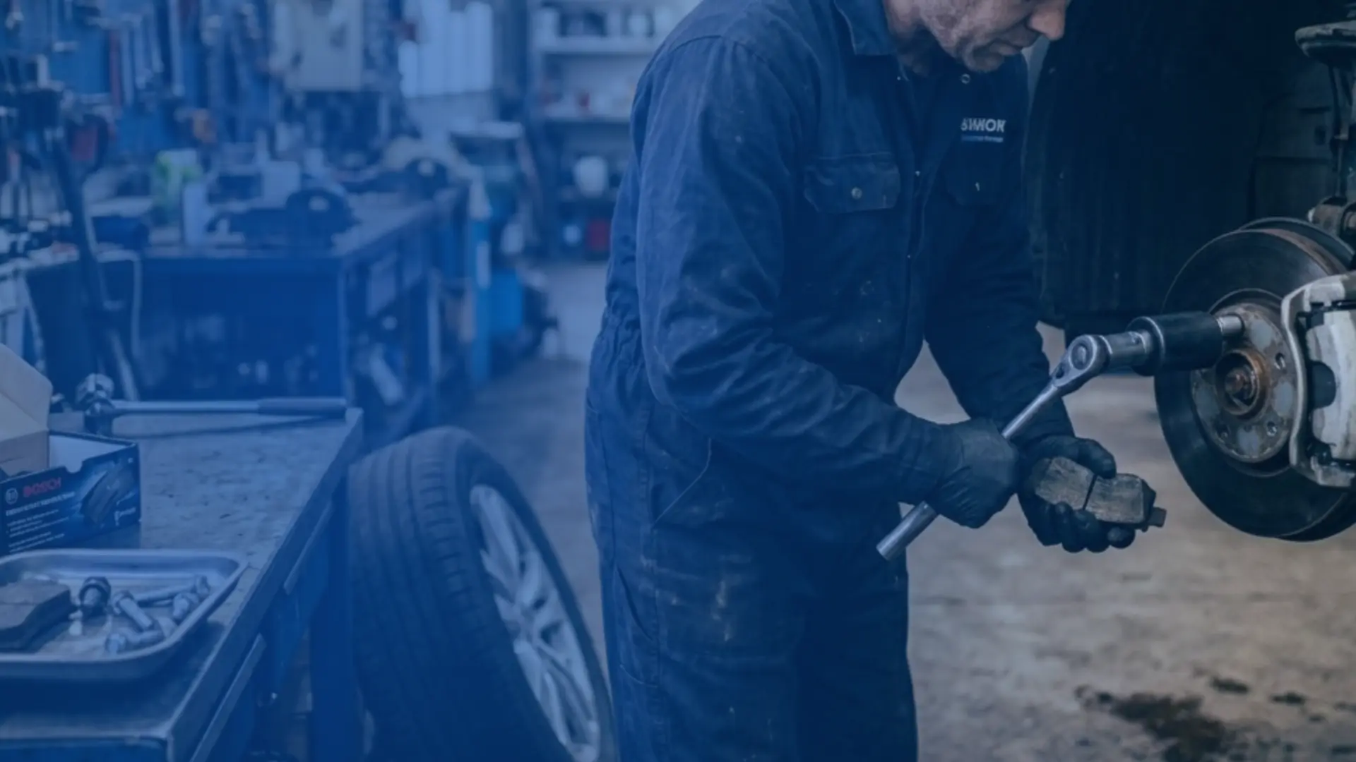 Mecânico com uniforme azul realizando manutenção preventiva no motor de um veículo com o capô aberto dentro de uma oficina mecânica.